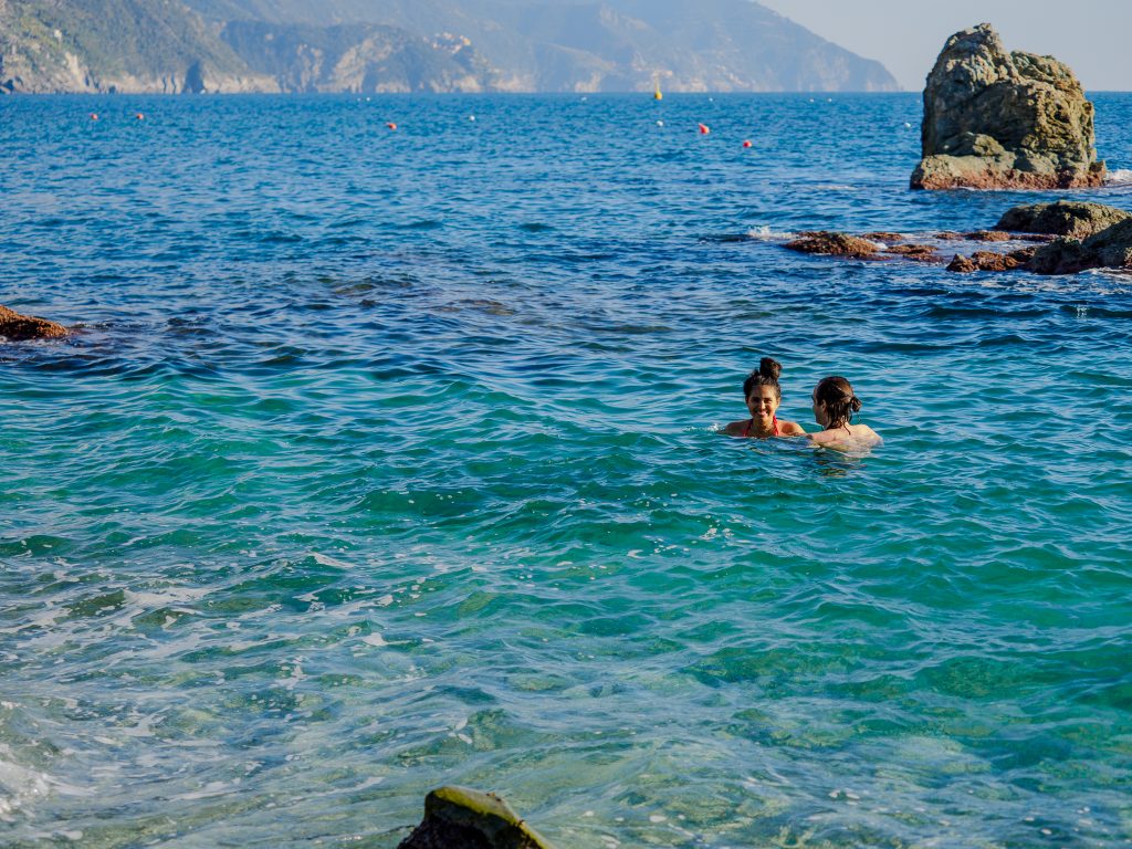 La costa di Monterosso con acqua turchese e speroni rocciosi; due persone nuotano nel mare vicino alle rocce, con scogliere lontane all'orizzonte.