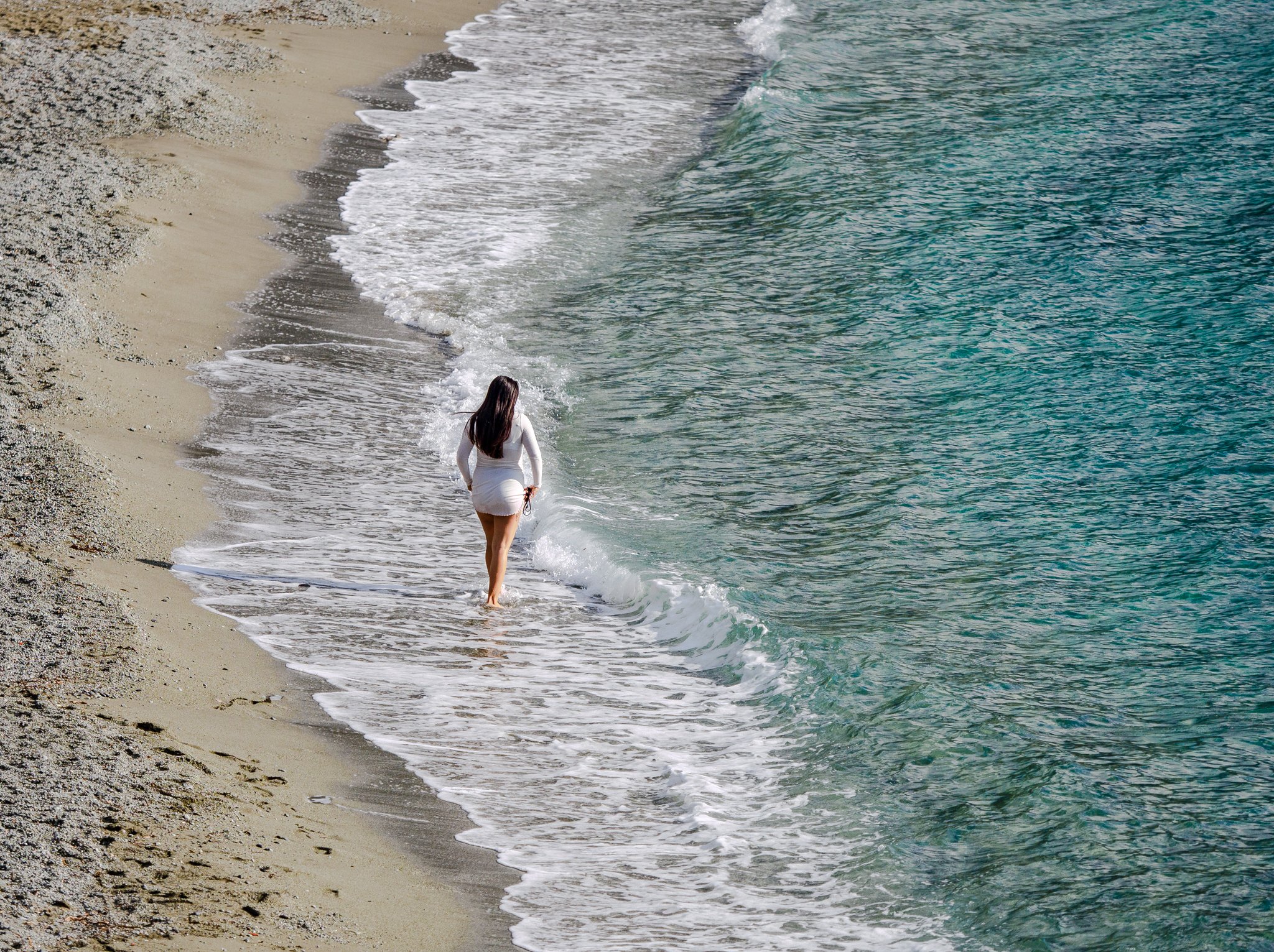 Mujer con vestido blanco caminando por la playa de Monterosso en Cinque Terre, con olas turquesas acariciando la costa de arena y un mar tranquilo bajo un cielo brillante.