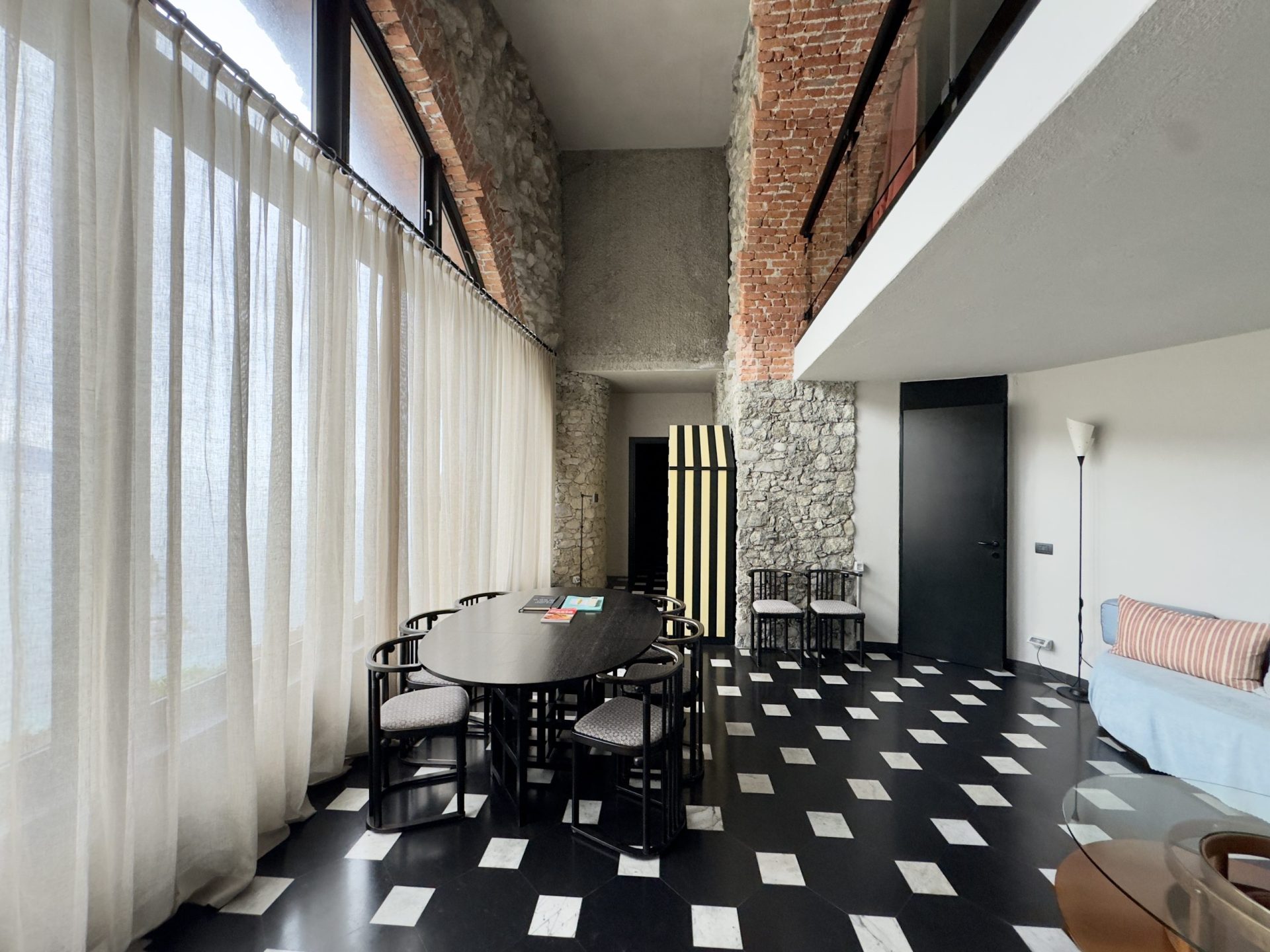 Interior of a modern living and dining area with a stone wall and brick elements, long sheer curtains along windows, a round black dining table with matching chairs, and a black-and-white checkered floor finishing with a sofa and floor lamp.