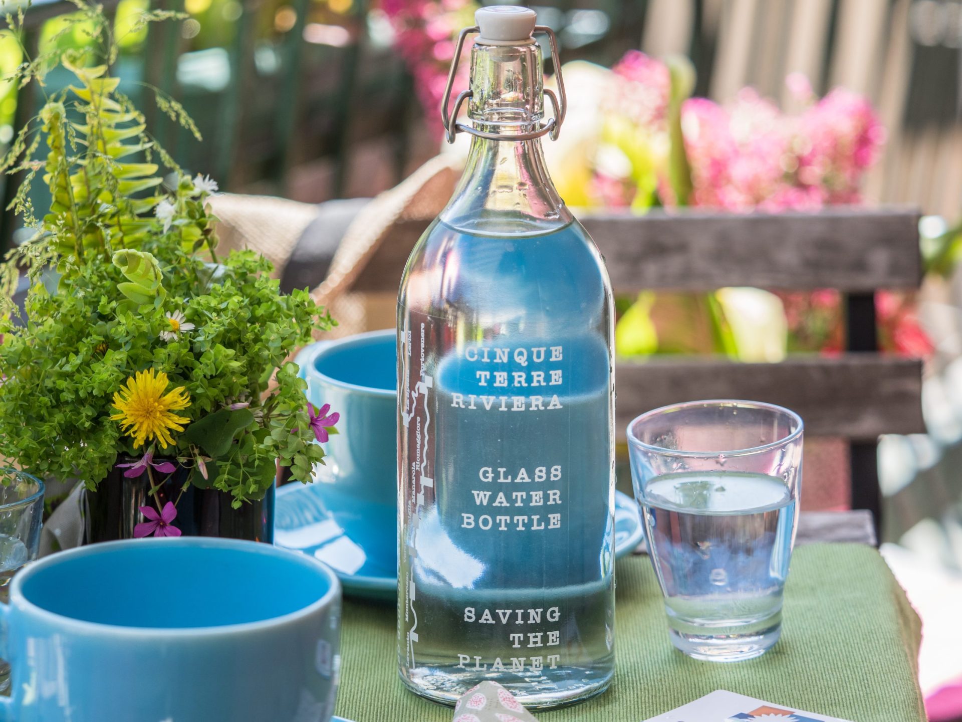 Glass water bottle labeled Cinque Terre Riviera on a table with blue cups and a glass of water, surrounded by potted greenery and colorful flowers outdoors.