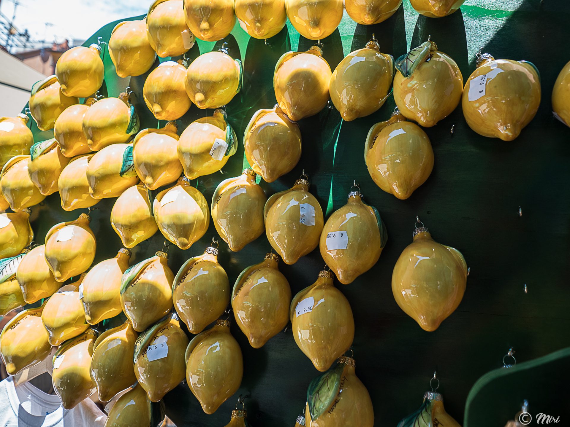 Racimo de adornos brillantes en forma de limón amarillo colgando de una pantalla verde con pequeñas etiquetas de precio, capturado a plena luz del día en un mercado costero.
