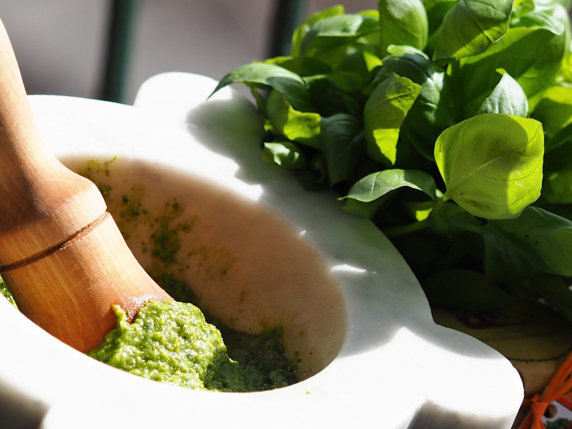 Fresco pesto verde pestato in un mortaio di pietra bianca con un pestello di legno, circondato da vivaci foglie di basilico per una preparazione di cucina italiana.