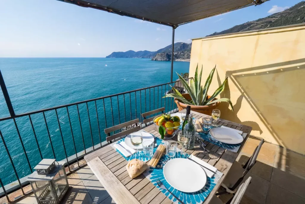 Vista sul mare blu da un balcone costiero con un tavolo da pranzo in legno apparecchiato per quattro, con una ciotola di frutta, pane, vino, piatti e una pianta di agave in vaso a Monterosso, vicino a La Casa del Gigante