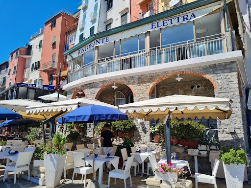 Pranzo all'aperto in un ristorante con facciata in pietra e portico ad archi, tavoli e sedie bianchi, ombrelloni blu e beige e vivace atmosfera mediterranea.