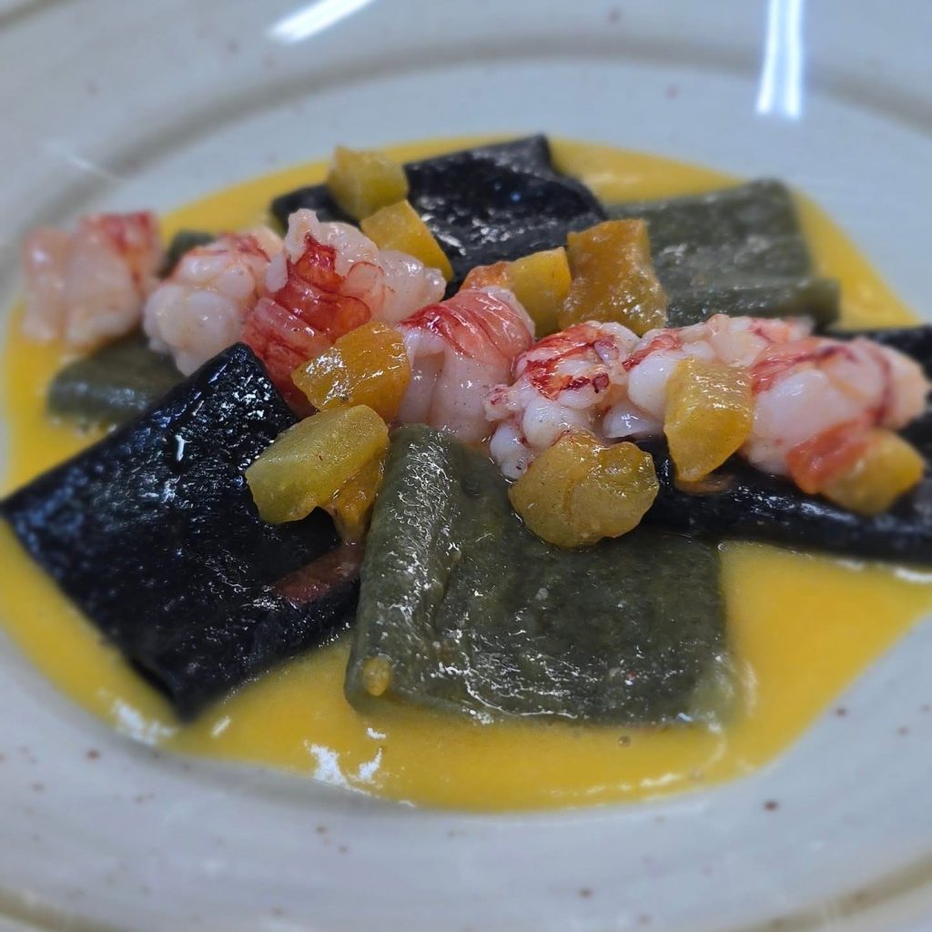 Plato de mariscos con camarones sobre pasta negra al tinta de calamar con acentos verdes y una vibrante salsa amarilla, presentado en un plato claro, cocina costera inspirada en Riomaggiore