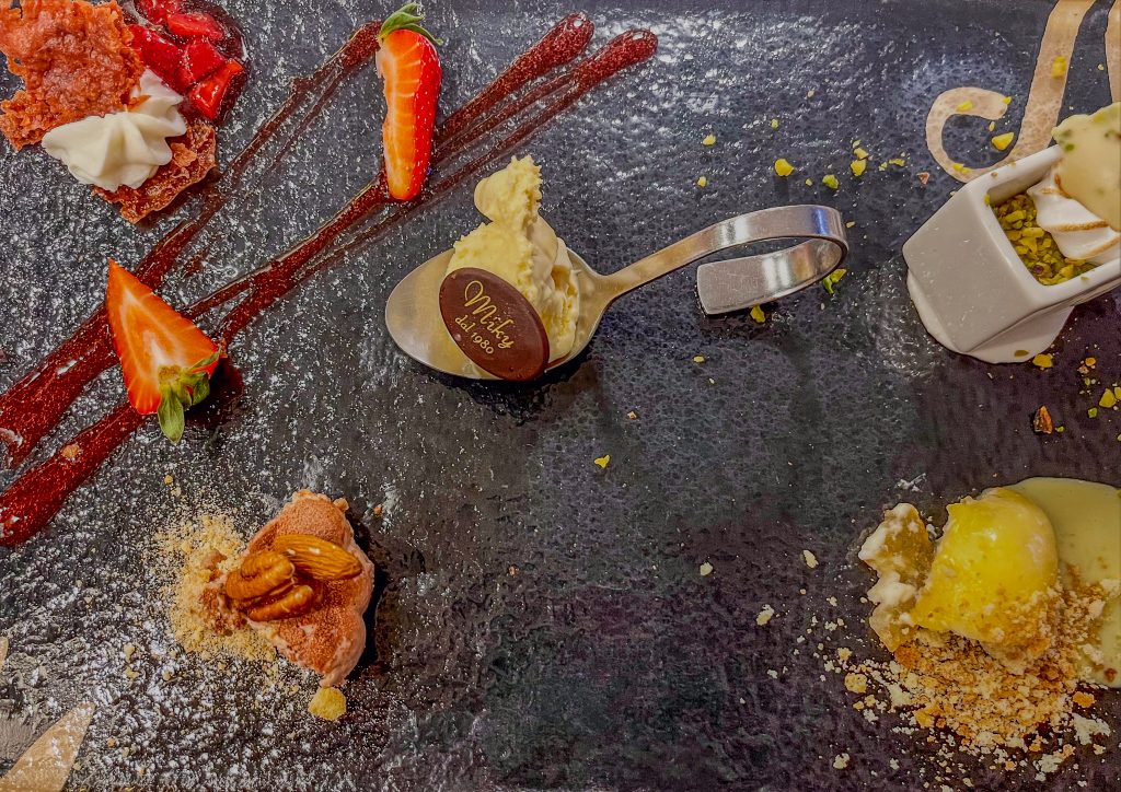 Assorted desserts and fresh fruit on a dark slate plate, featuring strawberries, dollops of cream, chocolate accents, and crumb toppings in an elegant tasting spread.