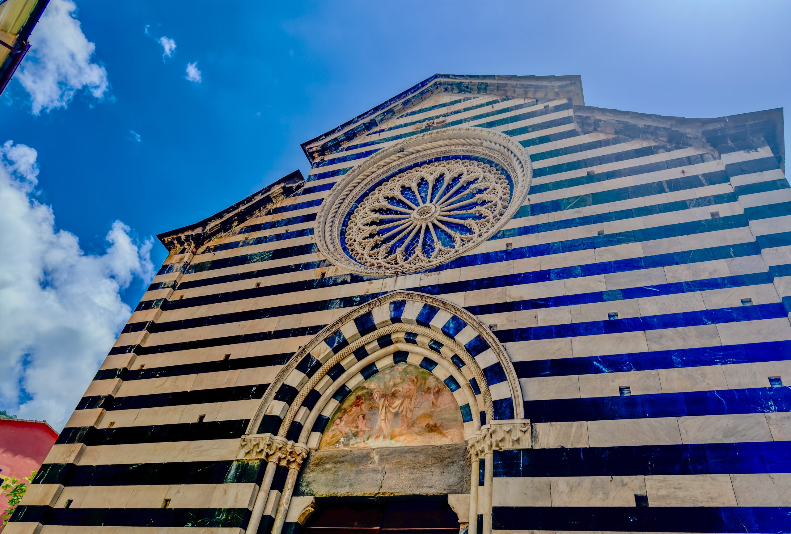 Facade of a church with bold blue and white horizontal stripes, a detailed circular rose window, and an arched doorway beneath a bright blue sky.
