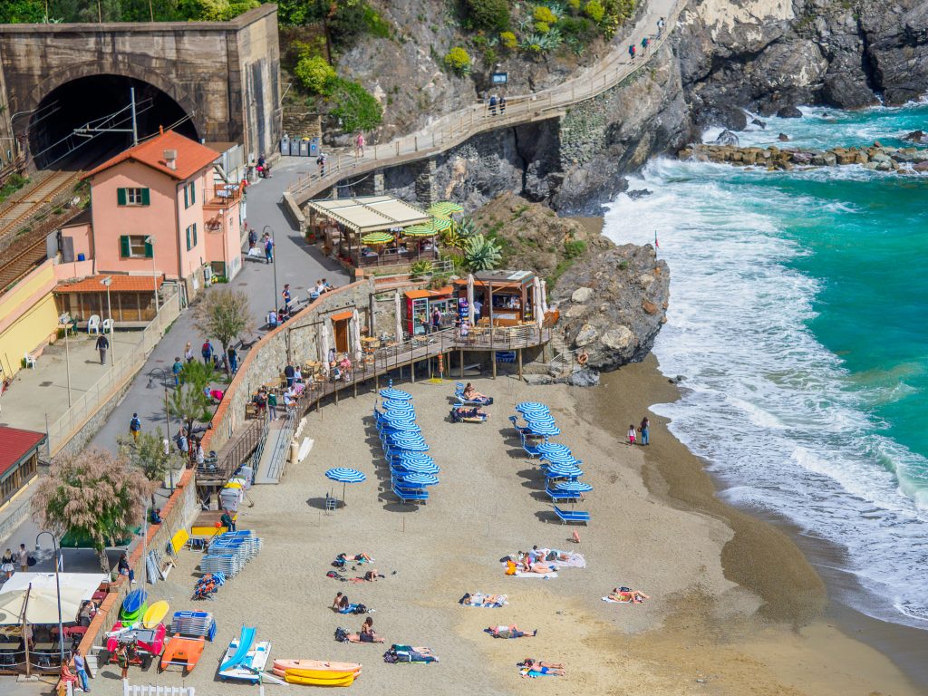 Aerial view of a rocky coastline with a sandy beach, turquoise sea, and blue striped umbrellas; colorful hillside buildings, a tunnel entrance, and a wooden promenade.