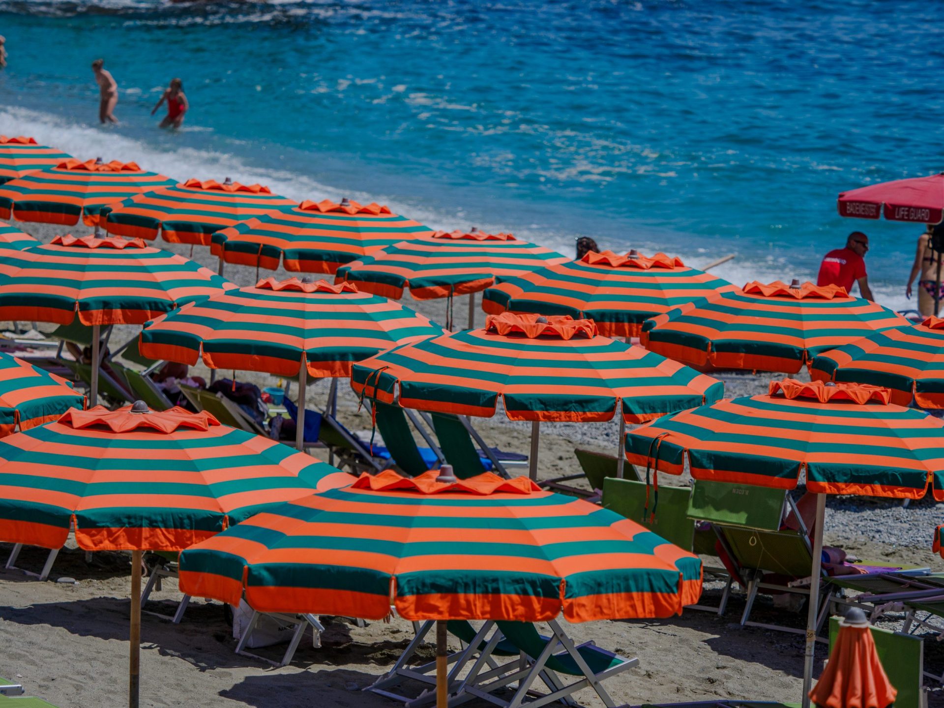 Hilera de sombrillas de playa a rayas naranjas y azul verdoso en una playa de arena con tumbonas, mar turquesa al fondo y algunas personas cerca del agua.
