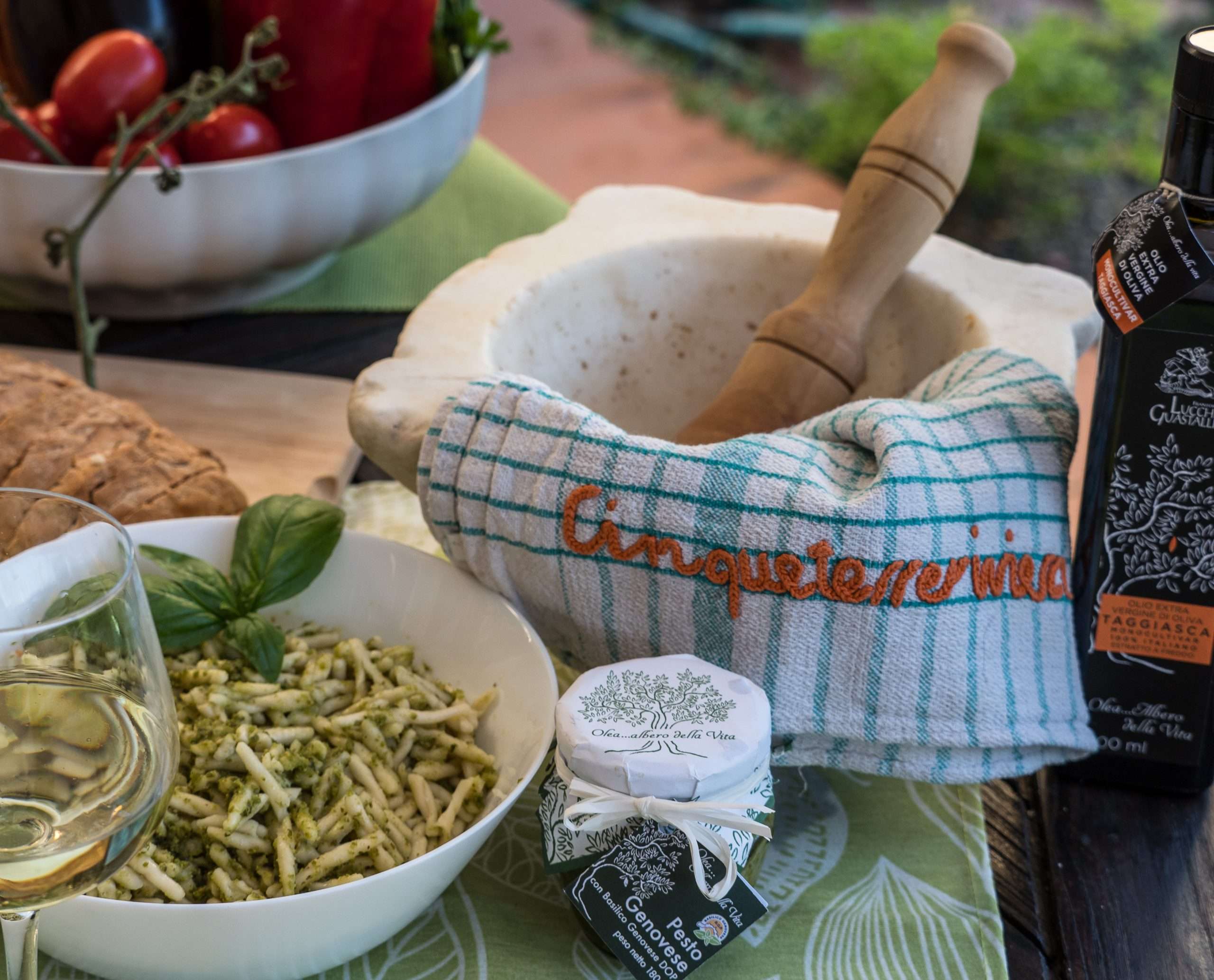Basilico fresco e pasta al pesto in una ciotola bianca, un mortaio e un pestello di legno con un asciugamano a righe, pomodorini e bottiglia di olio d'oliva su un tavolo scuro.