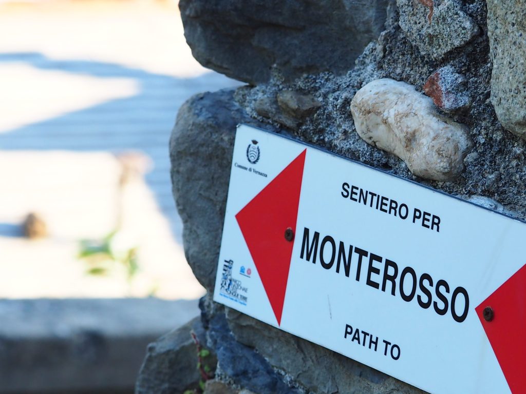 Monterosso path sign mounted on a rugged stone wall, featuring a bold red chevron and Italian text, captured along Cinque Terre Riviera’s coastal trail.