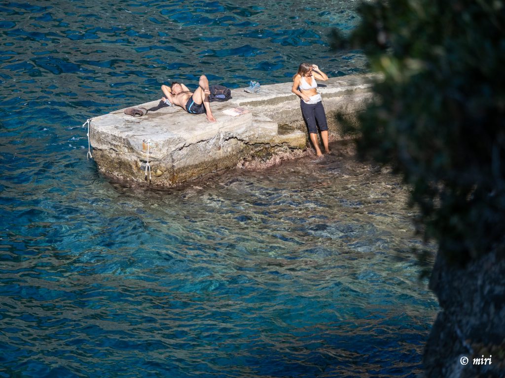 Dos personas tomando el sol en un muelle de hormigón desgastado que se adentra en aguas de color azul turquesa, una en bañador y la otra con una parte superior blanca y pantalones oscuros, con una costa rocosa cerca.