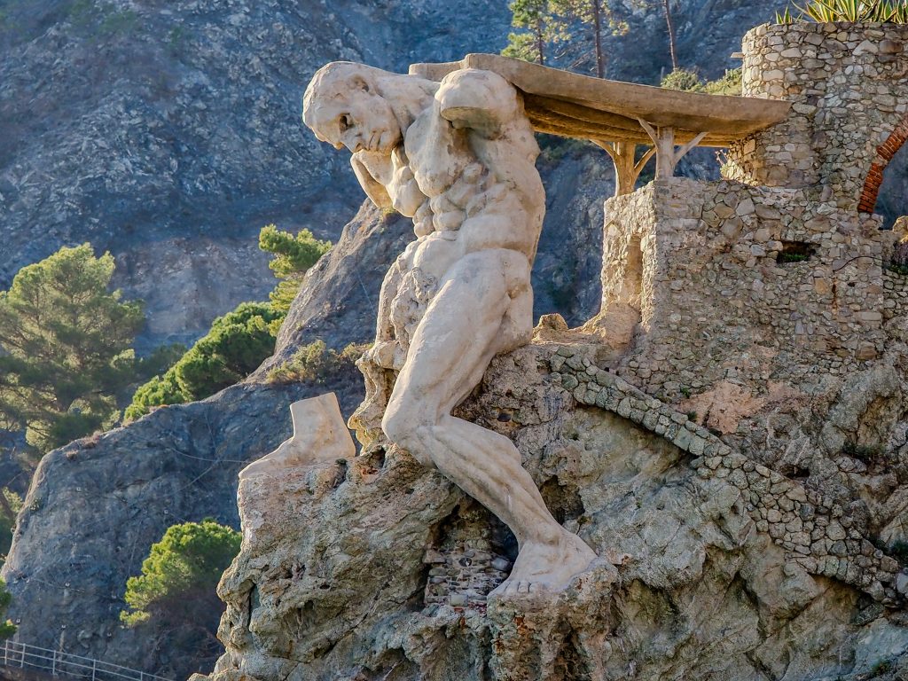 Stone statue of a muscular figure carrying a beam on a cliff, set among rocky terrain and a stone terrace with pine trees, showcasing weathered textures and dramatic coastal scenery.