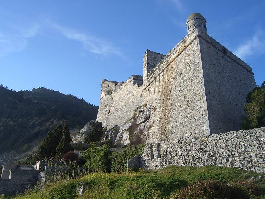 Il castello Doria di Portovenere con alte mura in pietra e una torre rotonda situato su un fianco di una collina rocciosa sotto un cielo azzurro e limpido, che mostra un terreno costiero accidentato nelle vicinanze