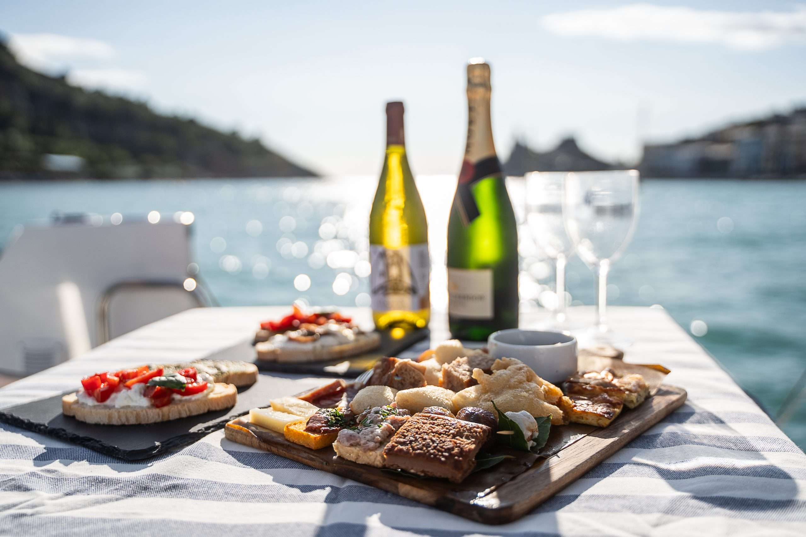 Esperienza enogastronomica Cinque Terre Riviera a bordo di una barca al tramonto: due bottiglie di champagne, bicchieri vuoti e un tagliere di legno con un assortimento di formaggi, pane e condimenti, sullo sfondo delle acque scintillanti della costa.