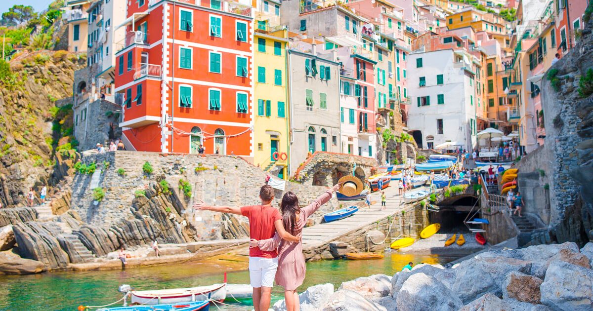 Colorato porto delle Cinque Terre con case pastolo lungo la banchina rocciosa, barche allineate e una coppia che si abbraccia godendosi la vista a Vernazza; tour in barca Grecale visibili nelle vicinanze