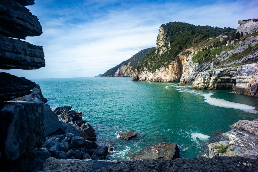 Il mare turchese che bagna le scoscese scogliere rocciose e la costa della Grotta di Byron nelle Cinque Terre, in Italia, caratterizzata da formazioni rocciose stratificate e da un cielo di un blu intenso.