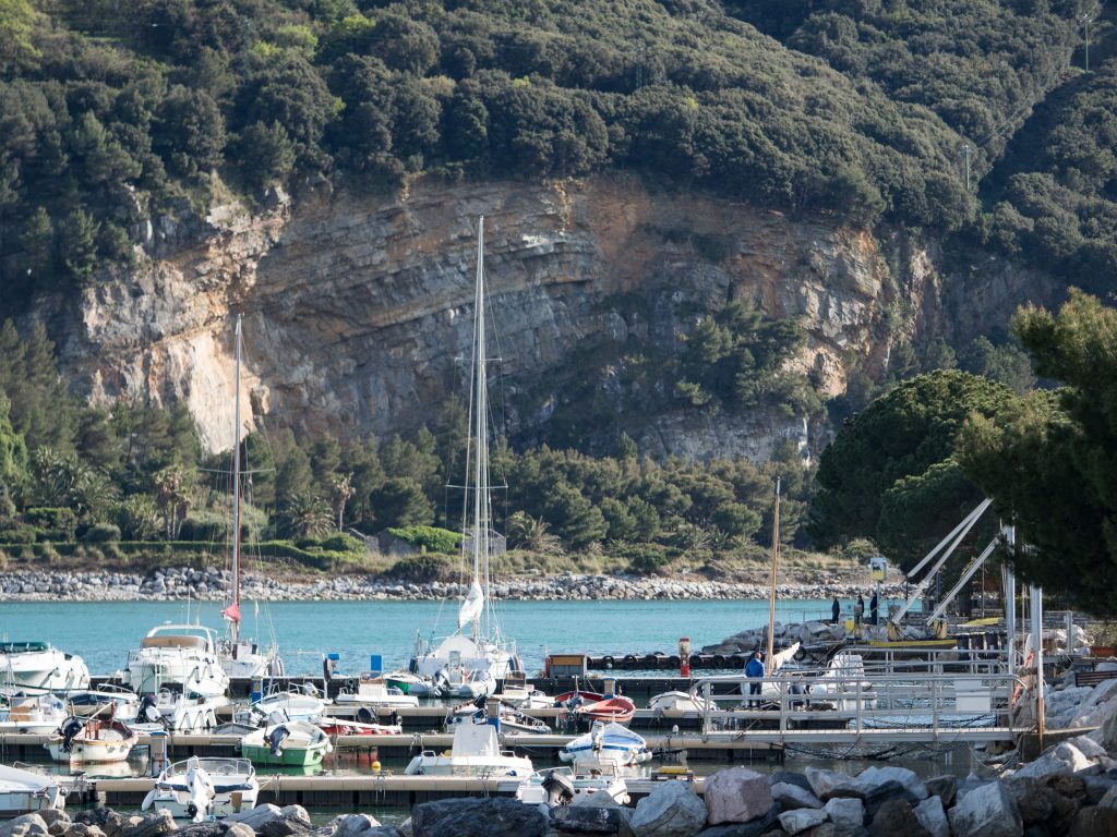 Scena costiera dell'Isola Palmaria con acqua turchese, un animato porticciolo di piccole barche e barche a vela, scogliere rocciose e alberi verdi lungo la costa