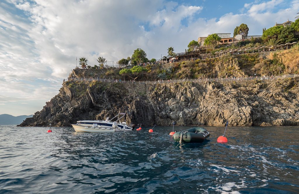 Costa rocosa de Cinque Terre con una ladera empinada, un sendero con barandilla, palmeras y barcos anclados en tranquilas aguas verde azuladas bajo un cielo nublado.