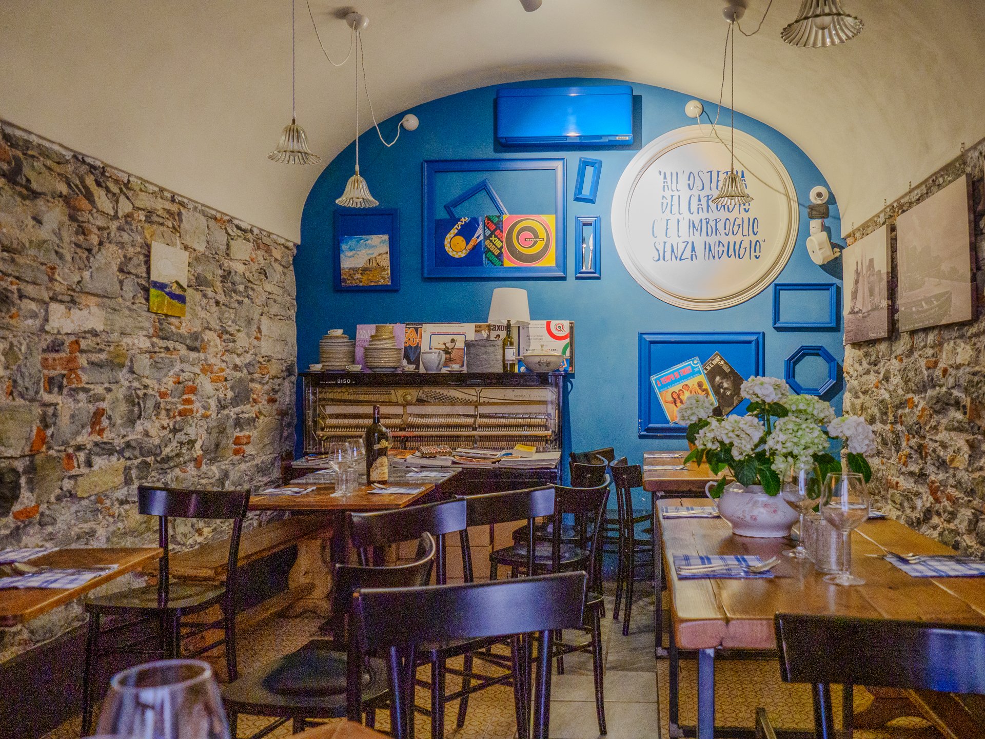 Interno dell'Osteria del Carugio con una parete curva blu brillante, lati in pietra a vista, tavoli da pranzo in legno, centrotavola floreale e arredamento vintage.