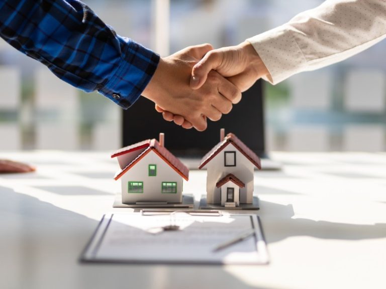 Un apretón de manos sobre unas casitas en miniatura con tejados rojos situadas en una mesa de planificación, con mangas azules y blancas, que representan a Cinque Terre Riviera Property Manager.