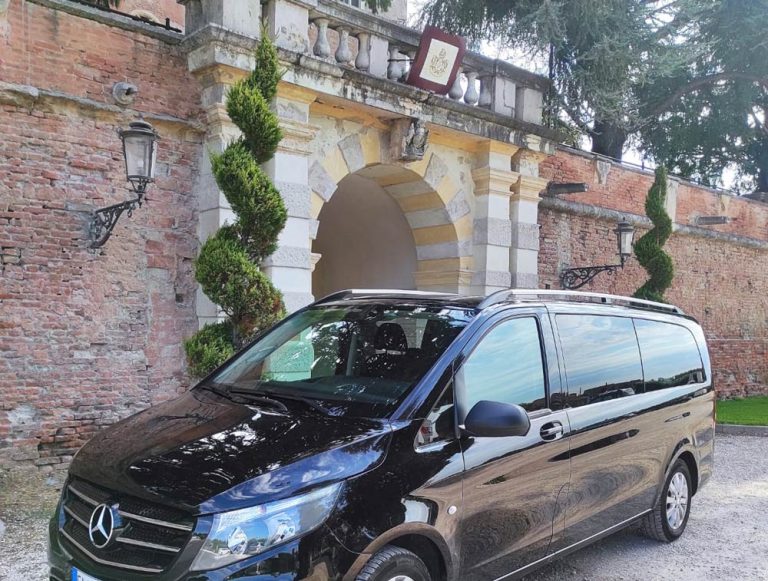 Un furgone Mercedes nero parcheggiato vicino a un antico arco in mattoni a Cinque Terre Riviera, con piante sempreverdi ben curate, classici muri in pietra e un’architettura storica sullo sfondo.