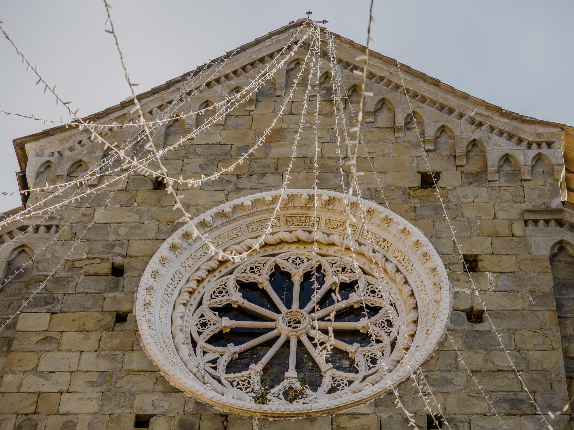 Corniglia church facade in Cinque Terre featuring a large circular rose window set in beige stone with decorative carvings and string lights draped across the facade.