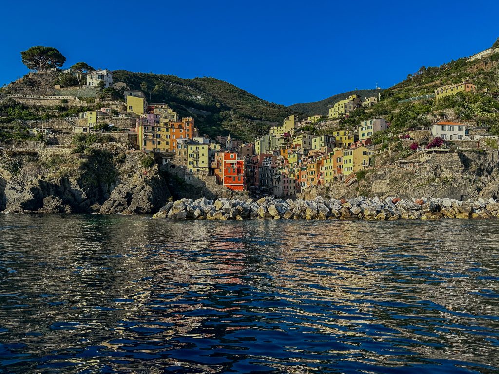 Pintoresco pueblo costero en ladera con casas pastel apiladas a lo largo de una costa rocosa bajo un cielo azul despejado, paisaje típico de Cinque Terre junto al agua.