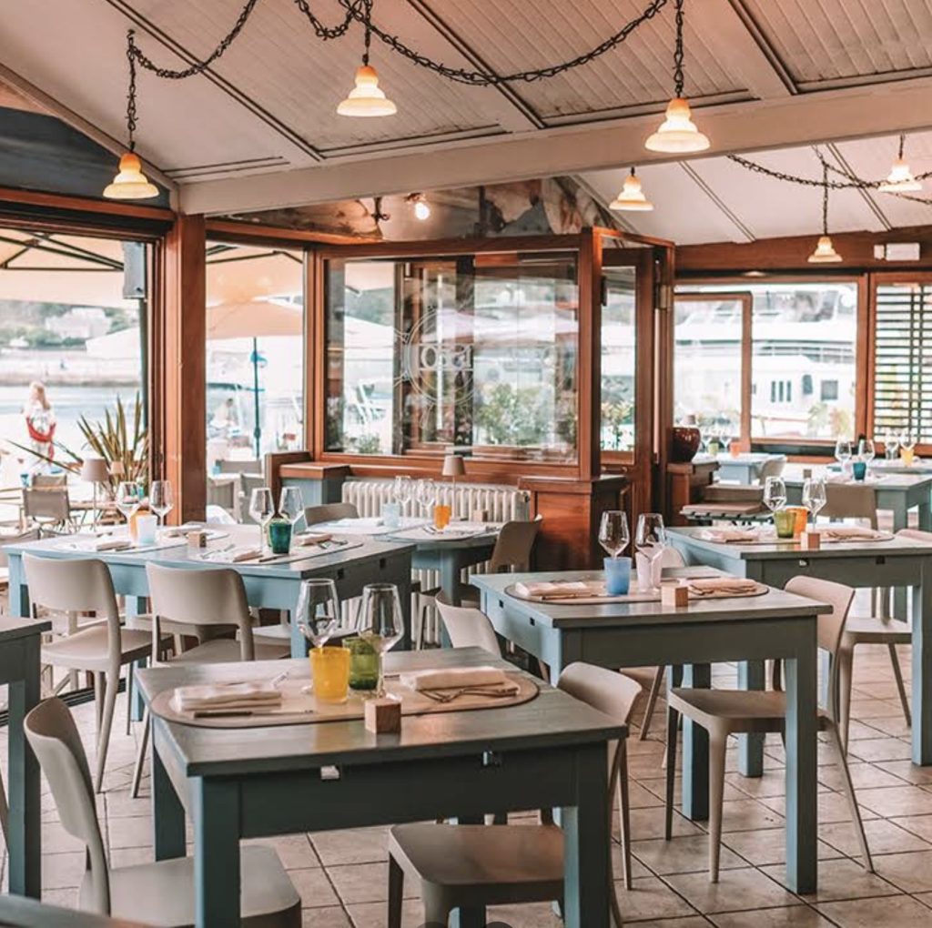 Interno di un ristorante sul mare con tavoli verde acqua, sedie bianche, bicchieri e calde luci a sospensione; grandi finestre rivelano una marina all'esterno.