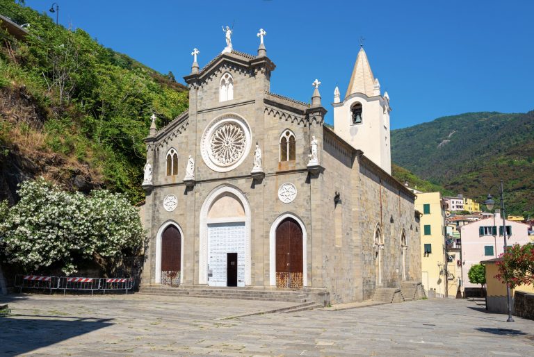 Fachada de iglesia de piedra con rosetón circular y dos puertas arqueadas gemelas, flanqueadas por estatuas, sobre un cielo azul brillante y ladera en Cinque Terre.
