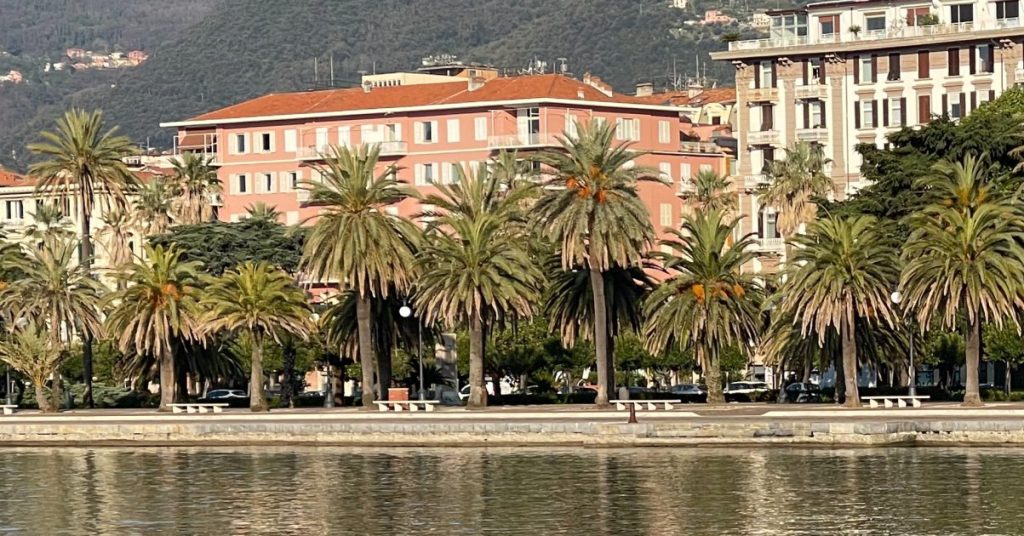 Hombre con chaqueta beige pescando desde una costa rocosa junto a un paseo marítimo bordeado de palmeras, con edificios de colores y colinas verdes al fondo, Cinque Terre Riviera.