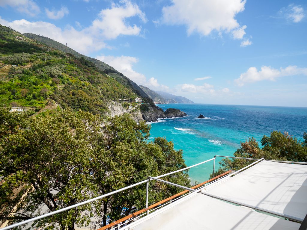 Vista costiera inondata di sole da una terrazza a Cinque Terre Riviera, con colline verdeggianti a terrazze, mare turchese, orizzonte blu e una ringhiera metallica in primo piano.