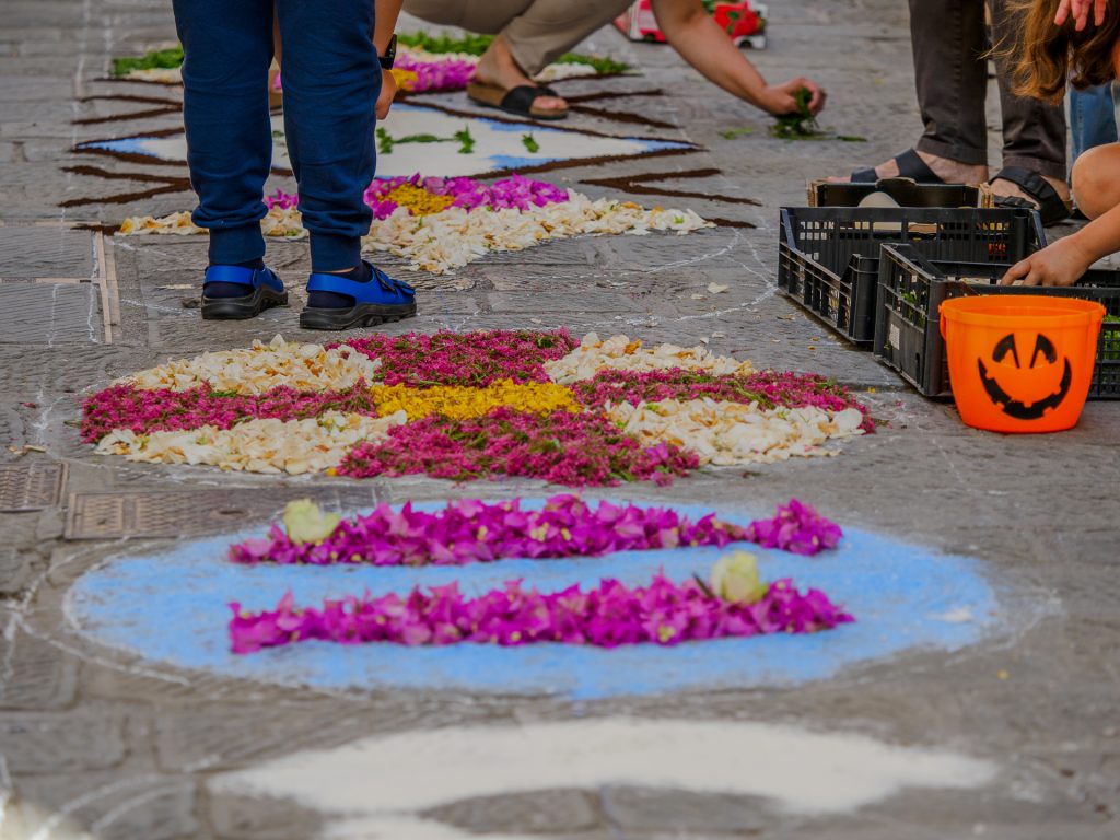 Colorful flower petals arranged on a gray stone street, with a bright orange Halloween pumpkin bucket and hands of people nearby, suggesting a sidewalk project or festival.
