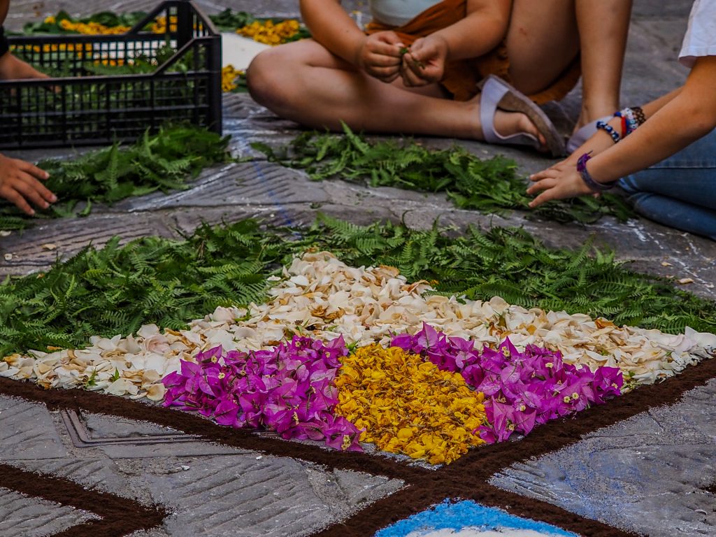 Colorful flower petals arranged in a decorative pattern on stone pavement, green ferns around the edges, and people seated nearby, suggesting a communal outdoor craft activity.