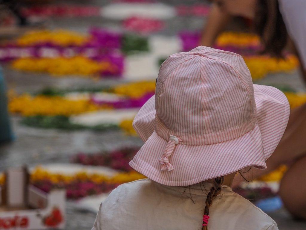 A child wearing a pink striped sunhat with a braid stands in front of a colorful outdoor flower market, soft-focus background with yellow and purple blooms.