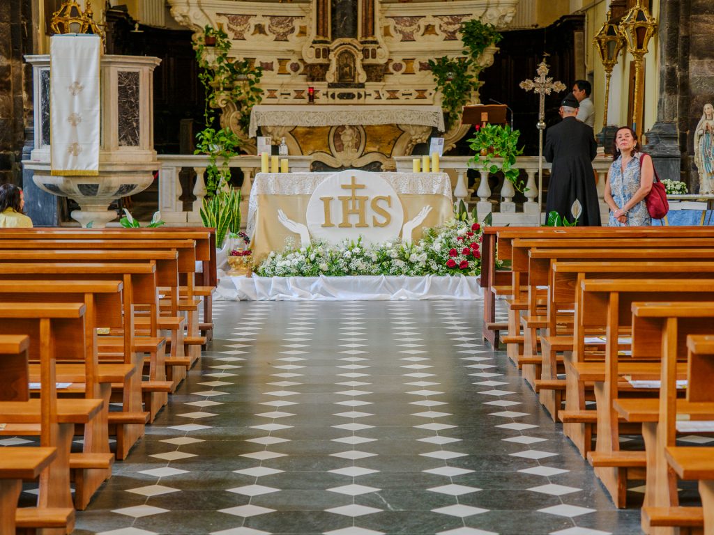 Interior of a church featuring an ornate altar with IHS symbol, white cloths, candles, and greenery, flanked by wooden pews and a checkered tile floor.