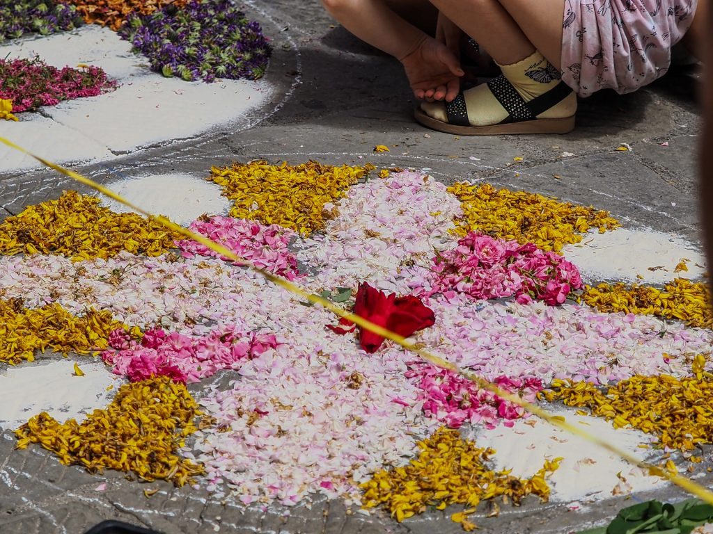Close-up view of pink and yellow flower petals arranged on a concrete pavement, with a child's foot in a sandal visible nearby, and a yellow string crossing the frame.