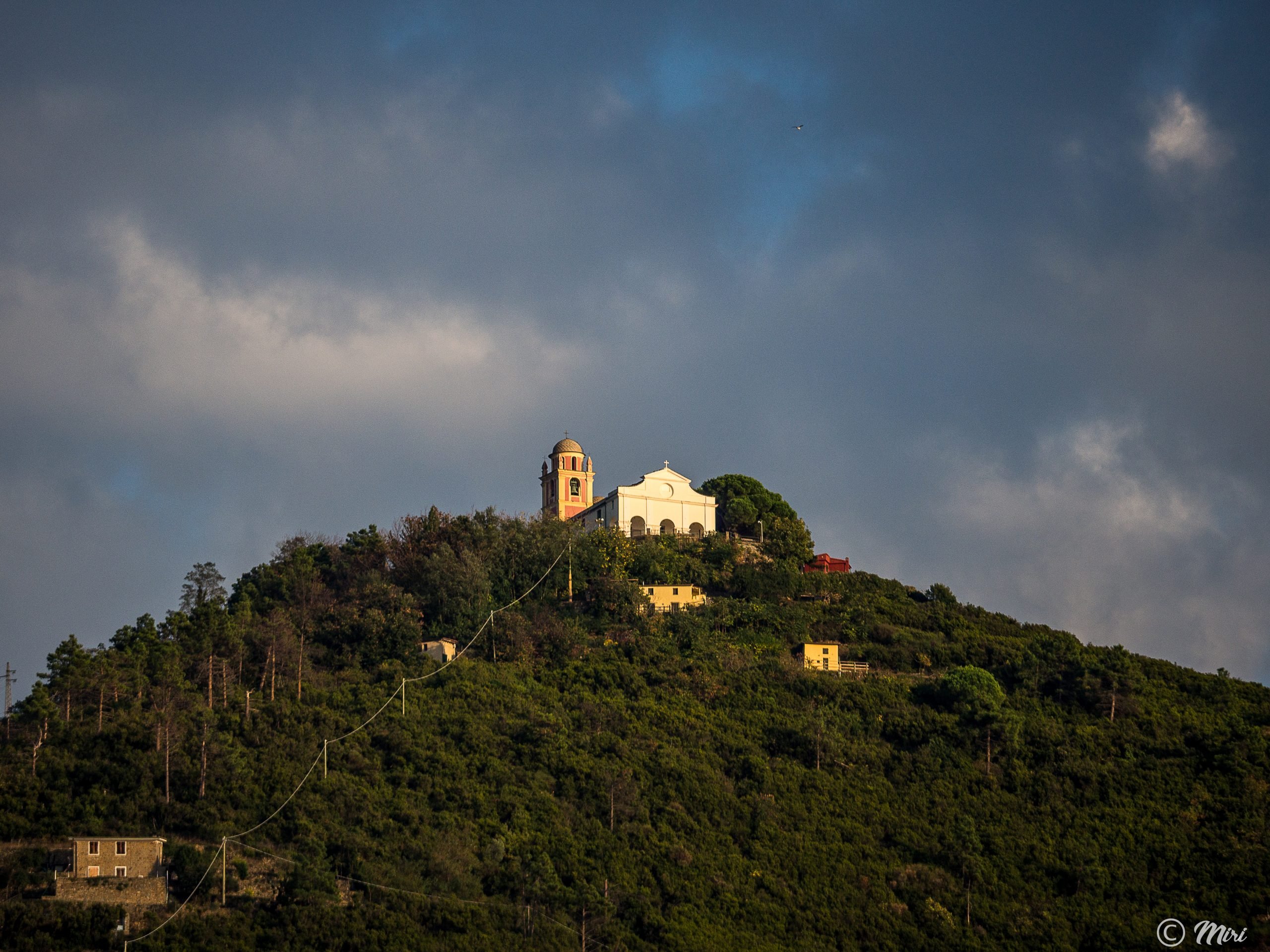 Santuario Montenero perched on a green hill overlooking Cinque Terre, a white-domed church framed by pine trees under a dramatic cloudy sky.