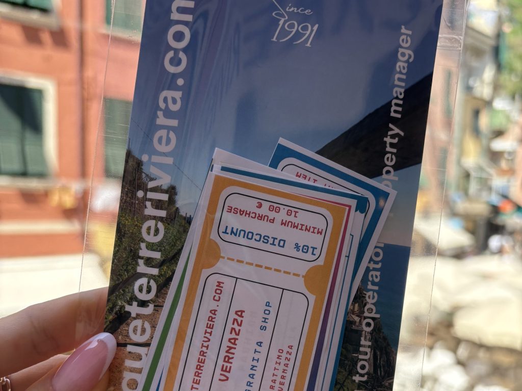 Un folleto azul y tarjetas coloridas sostenidas en una funda de plástico transparente, con un fondo de calle urbana y fachadas de edificios en la distancia.