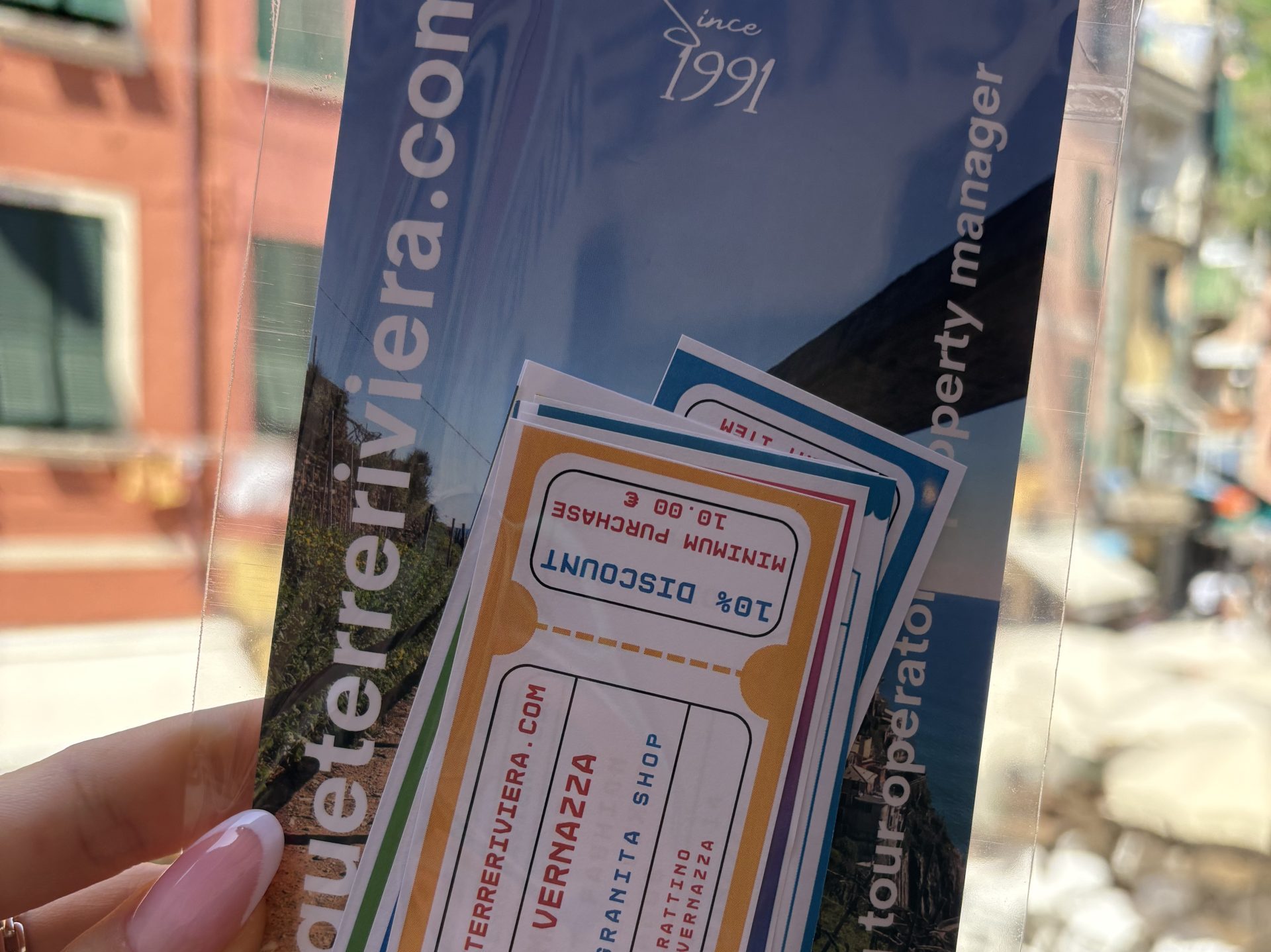 Un folleto azul y tarjetas coloridas sostenidas en una funda de plástico transparente, con un fondo de calle urbana y fachadas de edificios en la distancia.