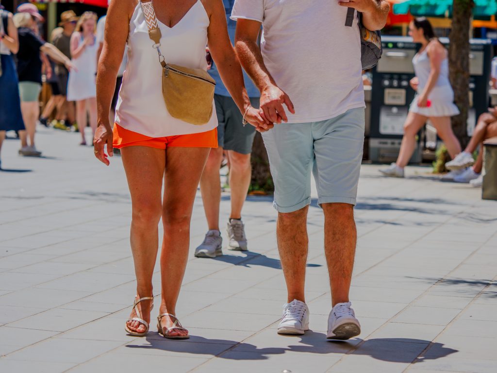 Una pareja camina por un paseo marítimo soleado en Vernazza, cogidos de la mano; la mujer lleva un top blanco sin mangas y pantalones cortos naranjas con un bolso bandolera bege, mientras que el hombre viste una camiseta blanca y pantalones cortos claros.