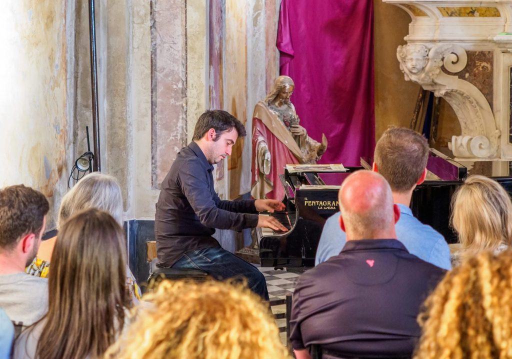 Un pianista con una camicia scura si esibisce a un pianoforte a coda con l'etichetta Il Pentagram La Spezia, in un interno storico con pareti in pietra, una statua e drappeggi cremisi, con il pubblico seduto nelle vicinanze.