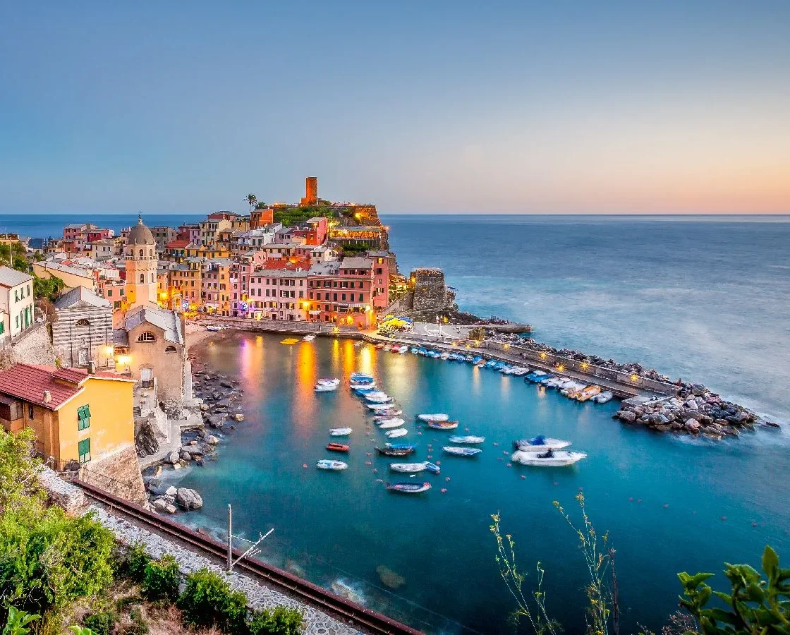 Il porto di Vernazza alle Cinque Terre al crepuscolo con colorati edifici collinari, piccole barche ancorate in acque turchesi e una tranquilla banchina costiera.