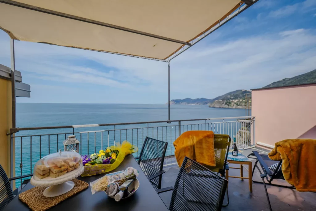 Terraza con balcón y vistas al mar azul en Cinque Terre, con una mesa de comedor preparada para aperitivos y bebidas, toallas naranjas colgadas en las sillas, una barandilla y colinas costeras distantes bajo un cielo despejado.