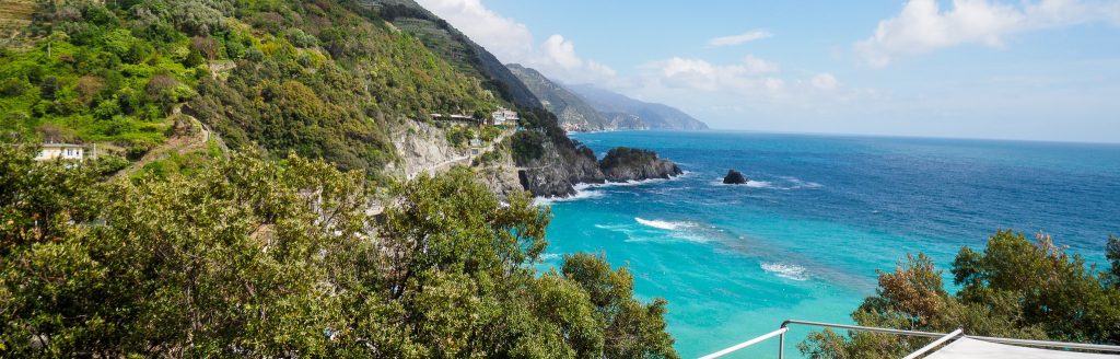 Vista desde la terraza de la costa de Cinque Terre con mar turquesa, colinas verde esmeralda, acantilados rocosos y un cielo azul brillante, perfecta para imágenes de alquiler vacacional.
