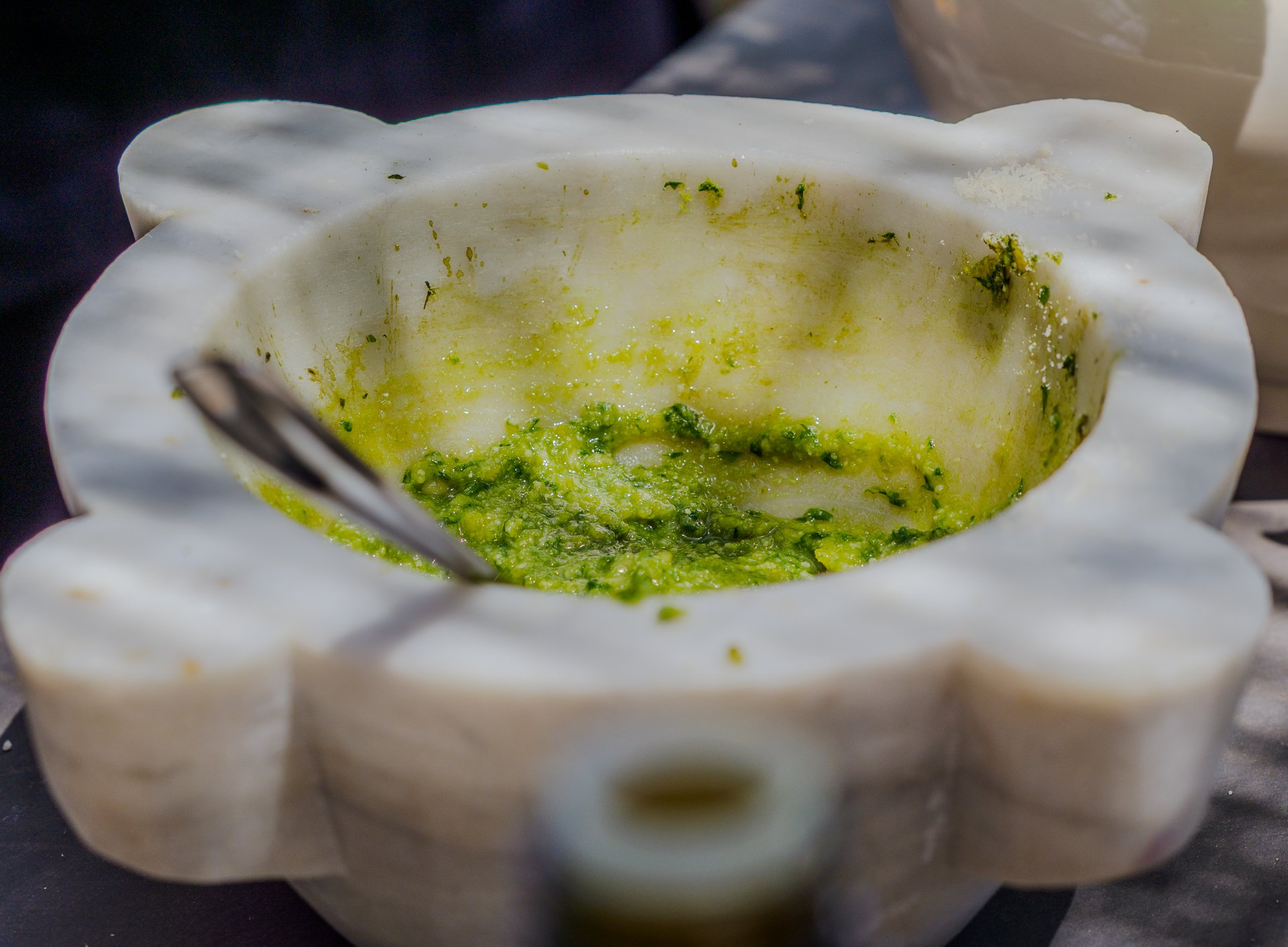 Mortero de mármol blanco con una mezcla de pesto verde y una cuchara dentro, colocado sobre una mesa oscura, evocando la escena culinaria de Monterosso en las Cinque Terre.