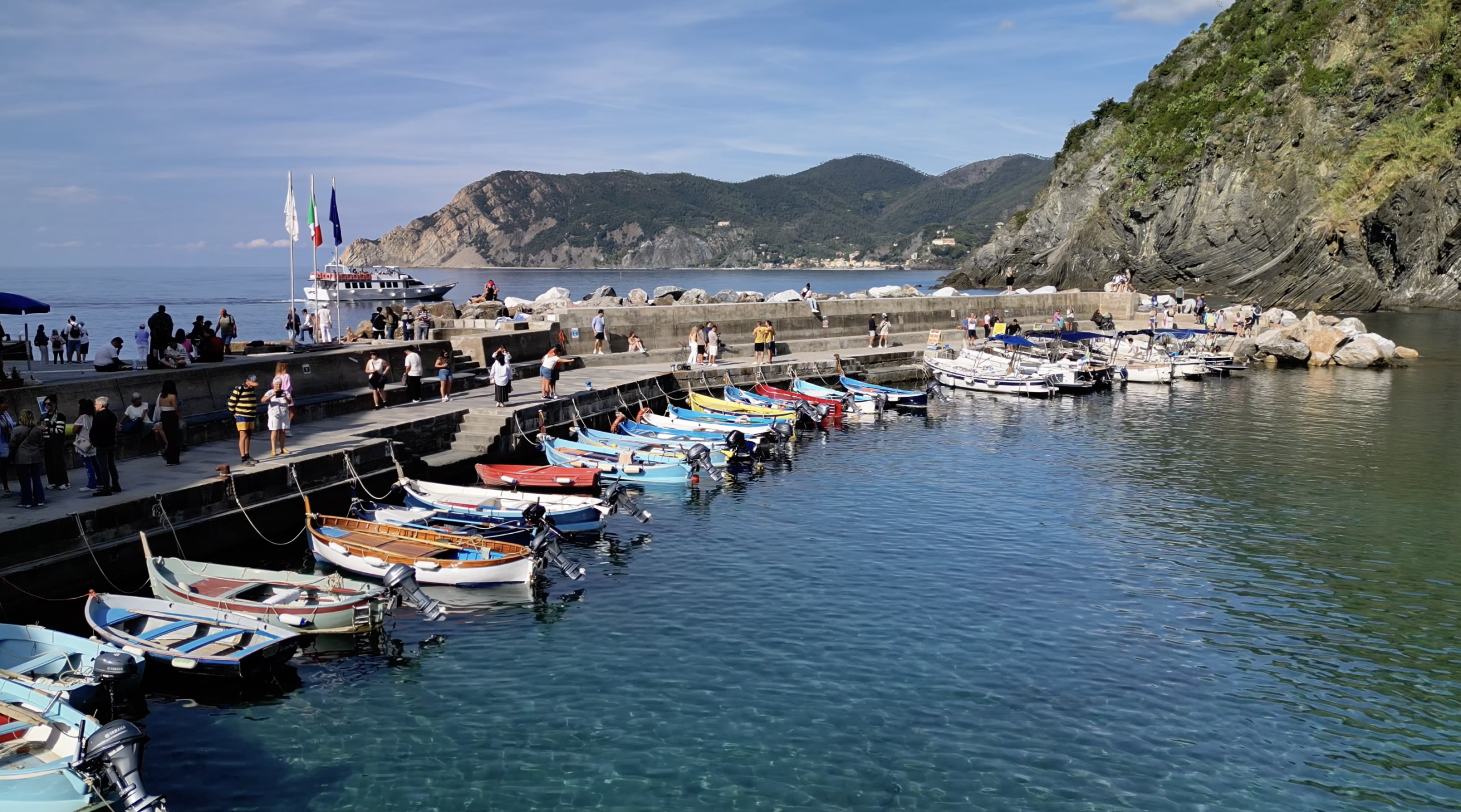 Pequeños barcos coloridos amarrados a lo largo de un puerto de piedra con agua azul cristalina, enmarcados por acantilados rocosos y un pueblo en la ladera de la colina en la región de Cinque Terre.