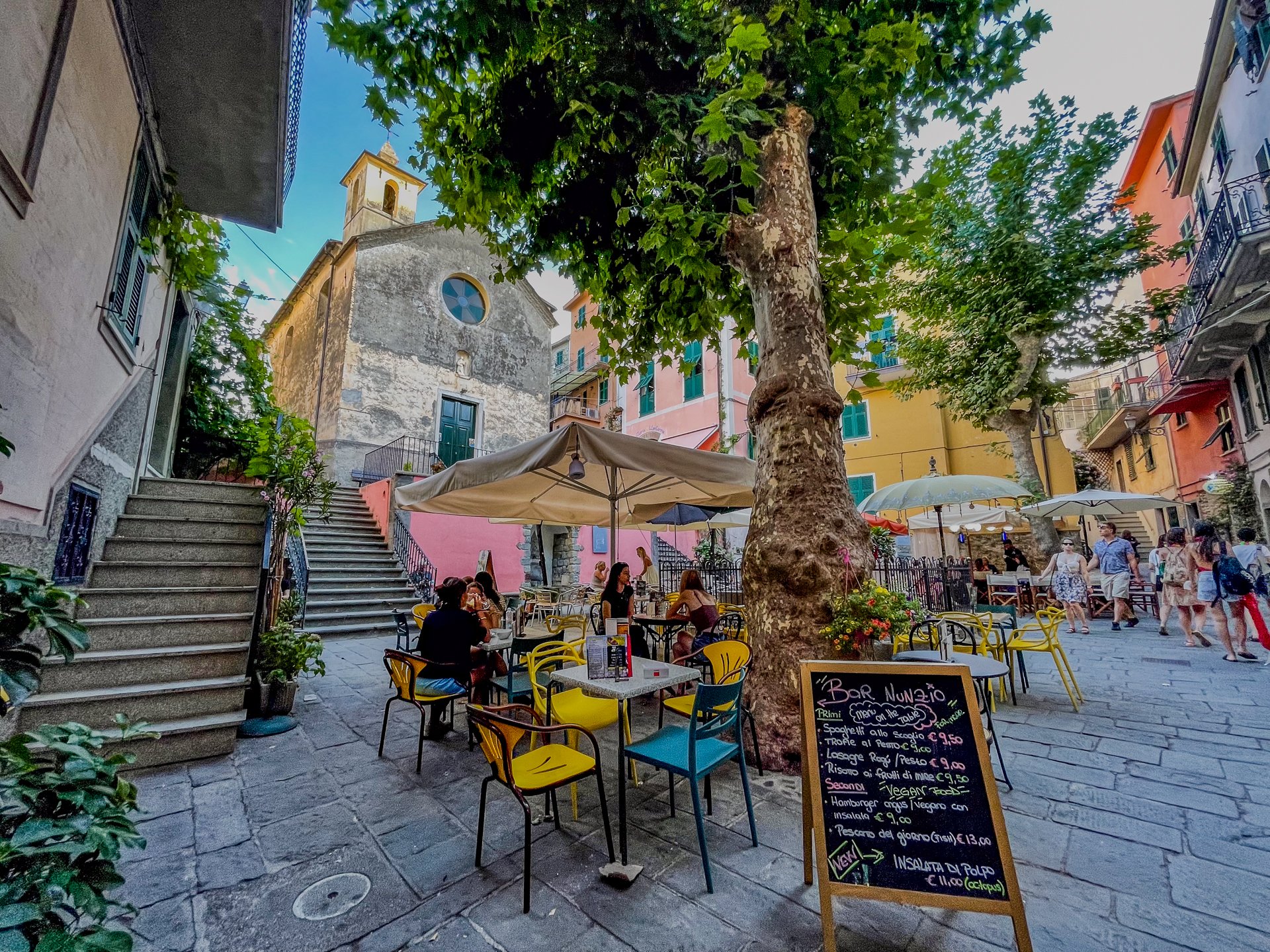 Plaza con cafeterías al aire libre en Corniglia, Cinque Terre, con edificios de colores, suelo de piedra, sillas amarillas y azules bajo las sombrillas y un gran árbol en el centro.