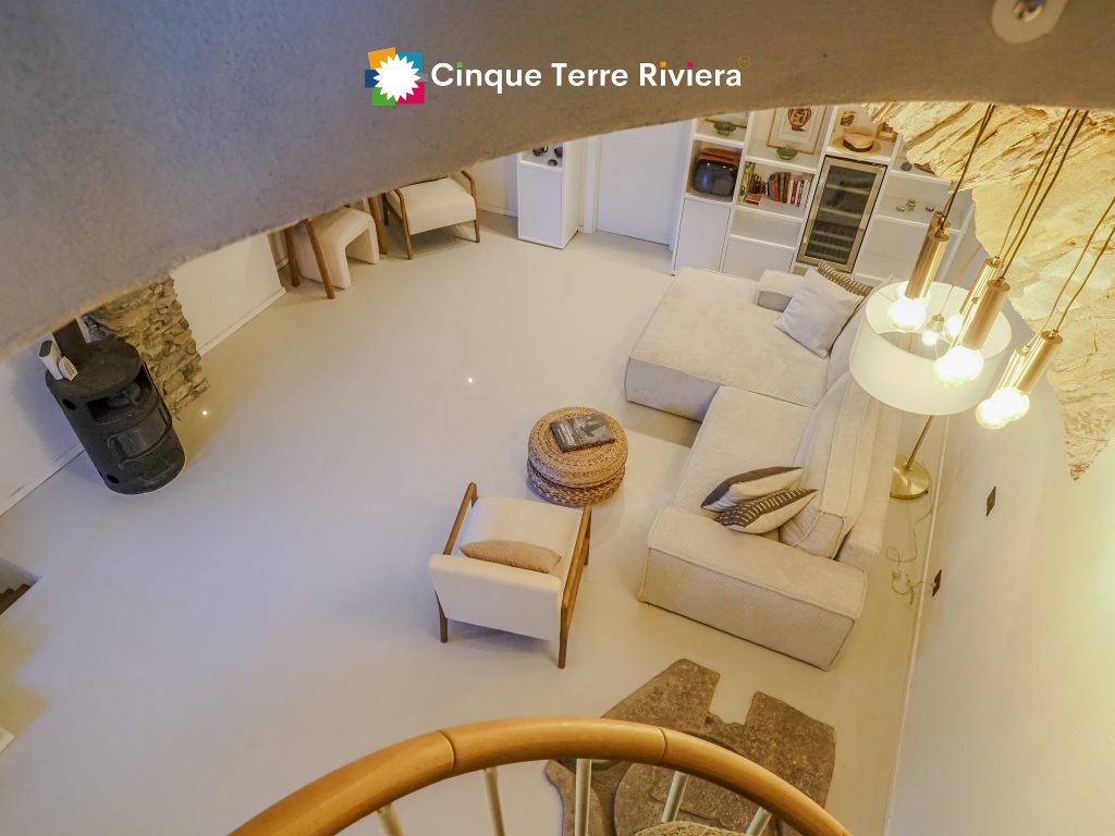 Interior de Frantoio: un salón rústico moderno en tonos beige en una casa de vacaciones de tipo Cinque Terre Riviera, con un sofá beige, paredes de piedra, detalles en madera y una cálida iluminación colgante.