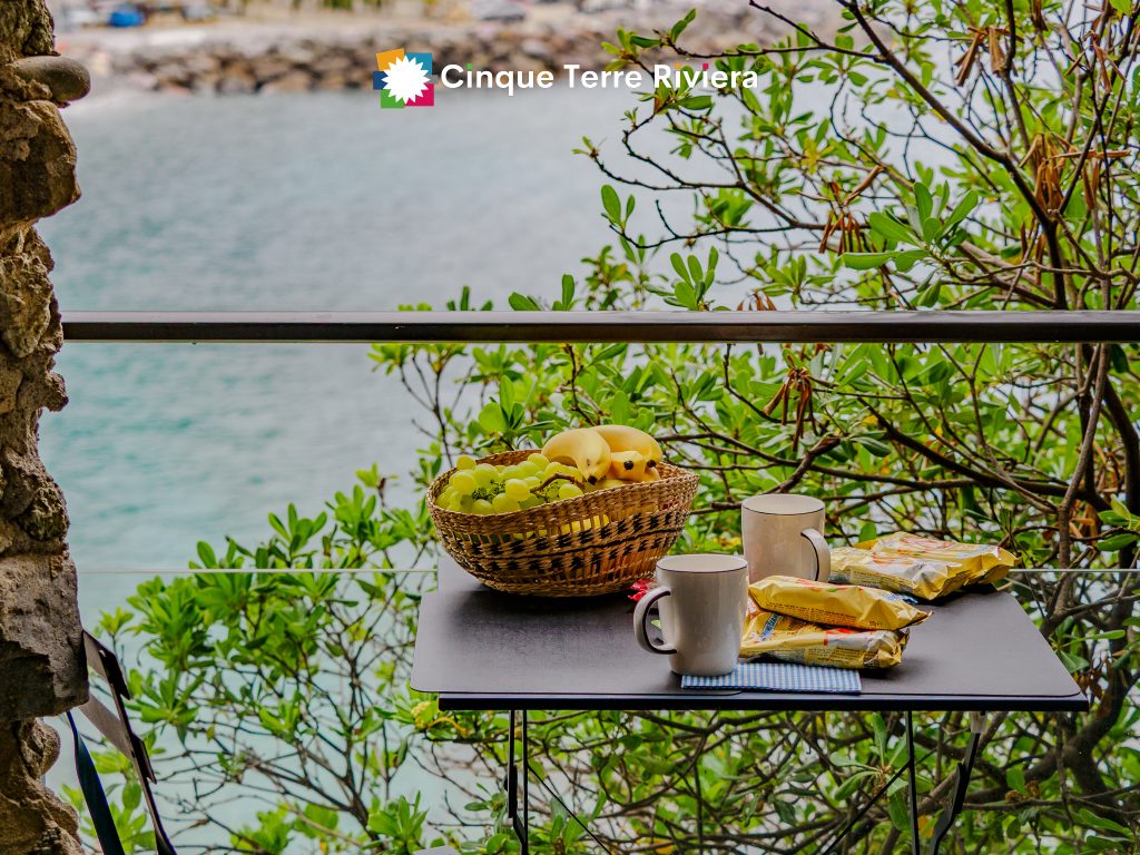 Mesa de balcón al aire libre con una cesta de mimbre que contiene uvas y plátanos, dos tazas, paquetes de aperitivos, plantas y vistas al mar; se aprecia el logotipo de Cinque Terre Riviera.